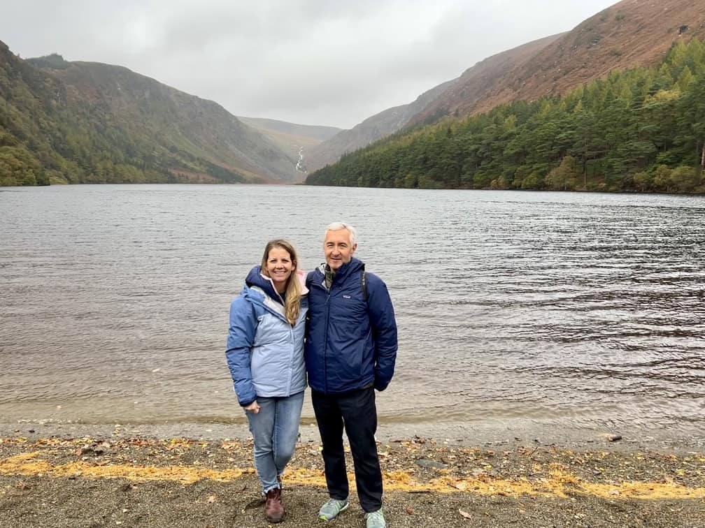 In this image a couple stands in front of the lake with the Wicklow Mountains in the background. The sky is grey and cloudy.