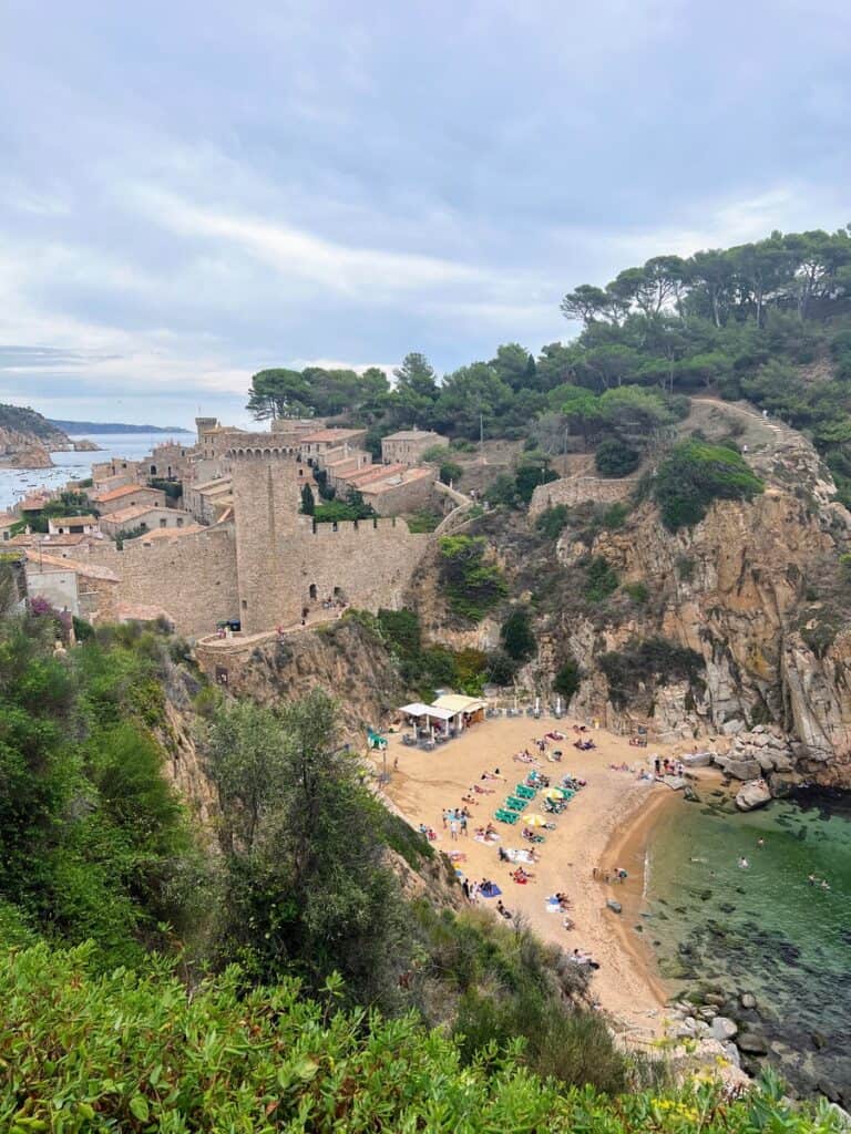 Es Codolar Beach with beach chairs and umbrellas with the medieval castle of Tossa de Mar just behind the beach. Crystal clear water in front of the beach.