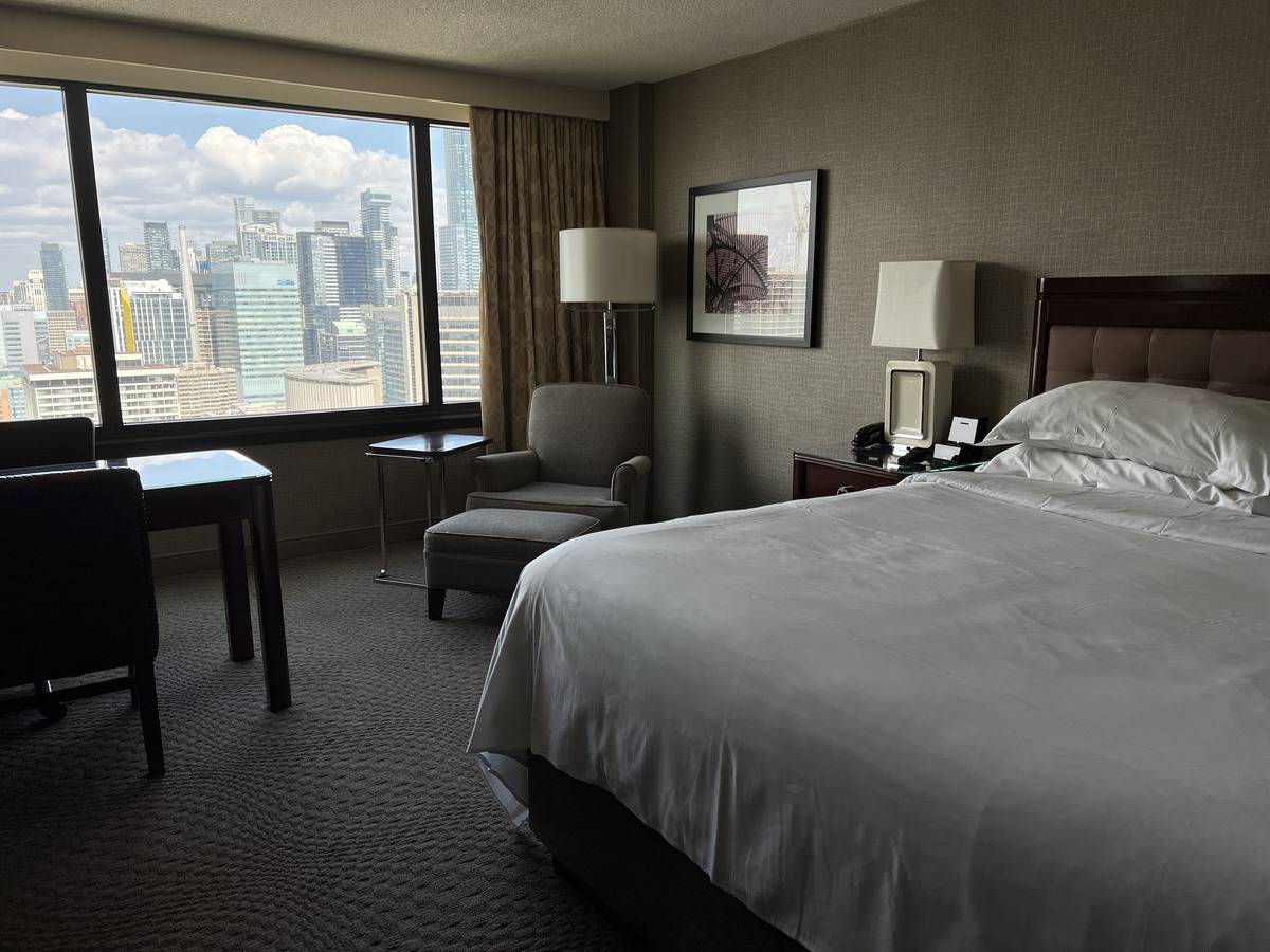 King size bed, desk, lounge chair, and view of downtown Toronto in a guest room of the Sheraton Centre Toronto Hotel Room.