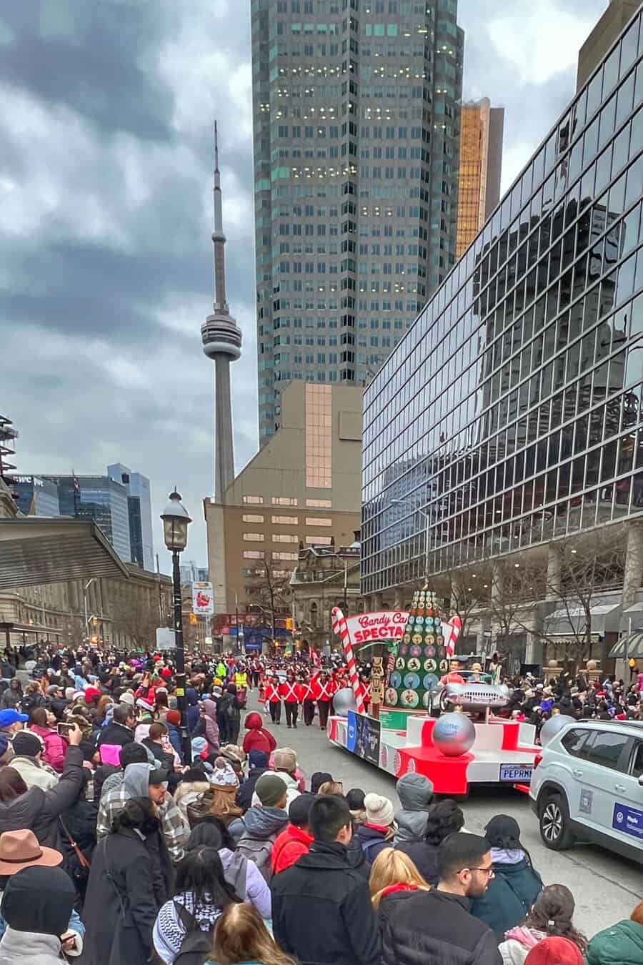Santa Claus Parade in downtown Toronto with large crowds lining the street to watch and the CN Tower visible in the background. A float with a Christmas Tree is visible in the parade with a marching band behind.