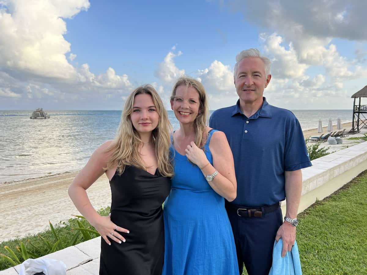 A smiling couple and their teenage daughter pose in front of the beach at Cancun Moon Place for a family photo.