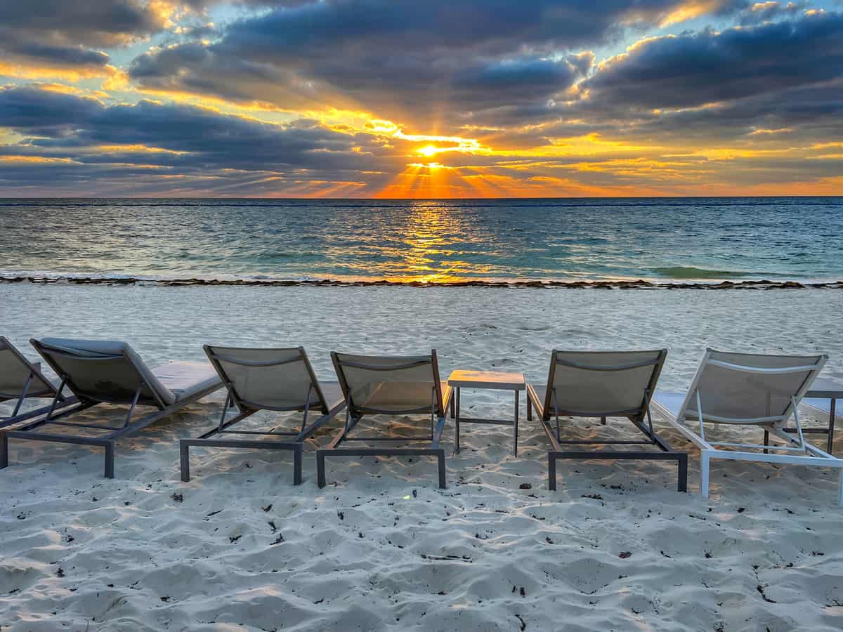 Empty Pool loungers overlook the beach at Moon Palace Cancun at sunrise. The sun peaks through the clouds above the Caribbean Sea.