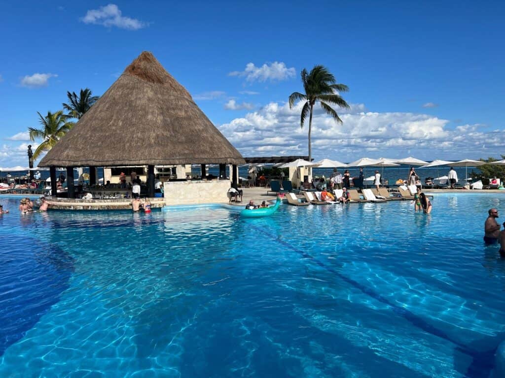 Large swimming pool with a few swimmers. Large swim-up bar is in the center of the pool and the sea can be seen in the distance behind the pool.