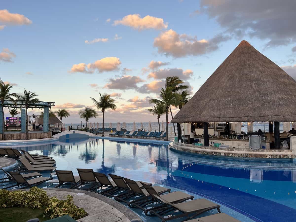 Swimming pool with chair loungers and a swim up bar with a thatched roof. A small bridge crosses over the pool and the beach can be seen in the distance.