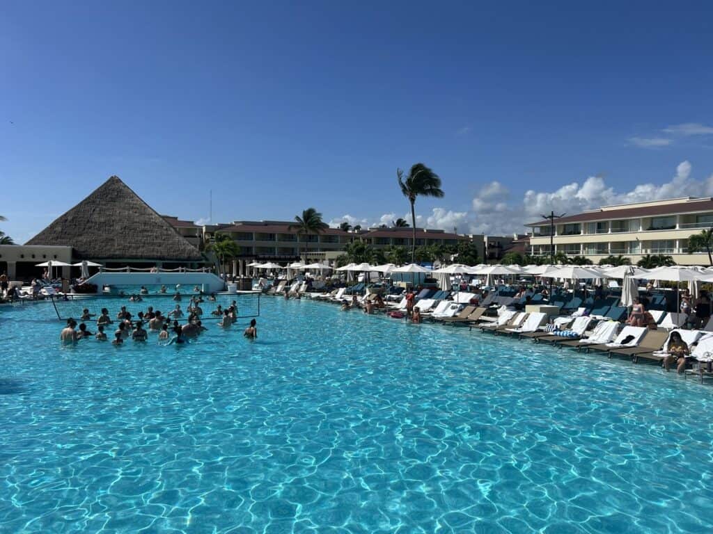 Resort guests play water volleyball in a very large pool. Chair loungers are set up on the edge of the pool.