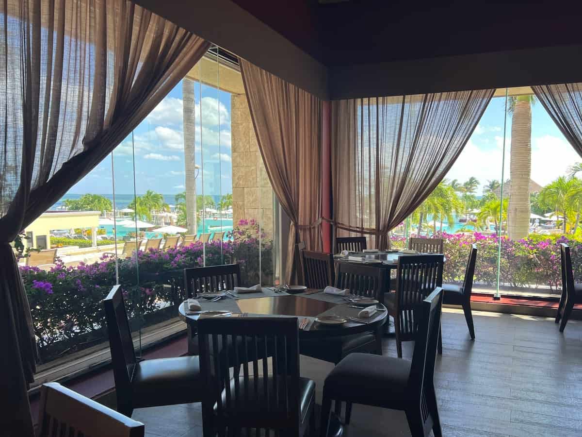 Round table with five chairs set up in front of floor to ceiling windows at a restaurant. Through the windows you can see the pool at Sunrise resorts in Cancun, Mexico.
