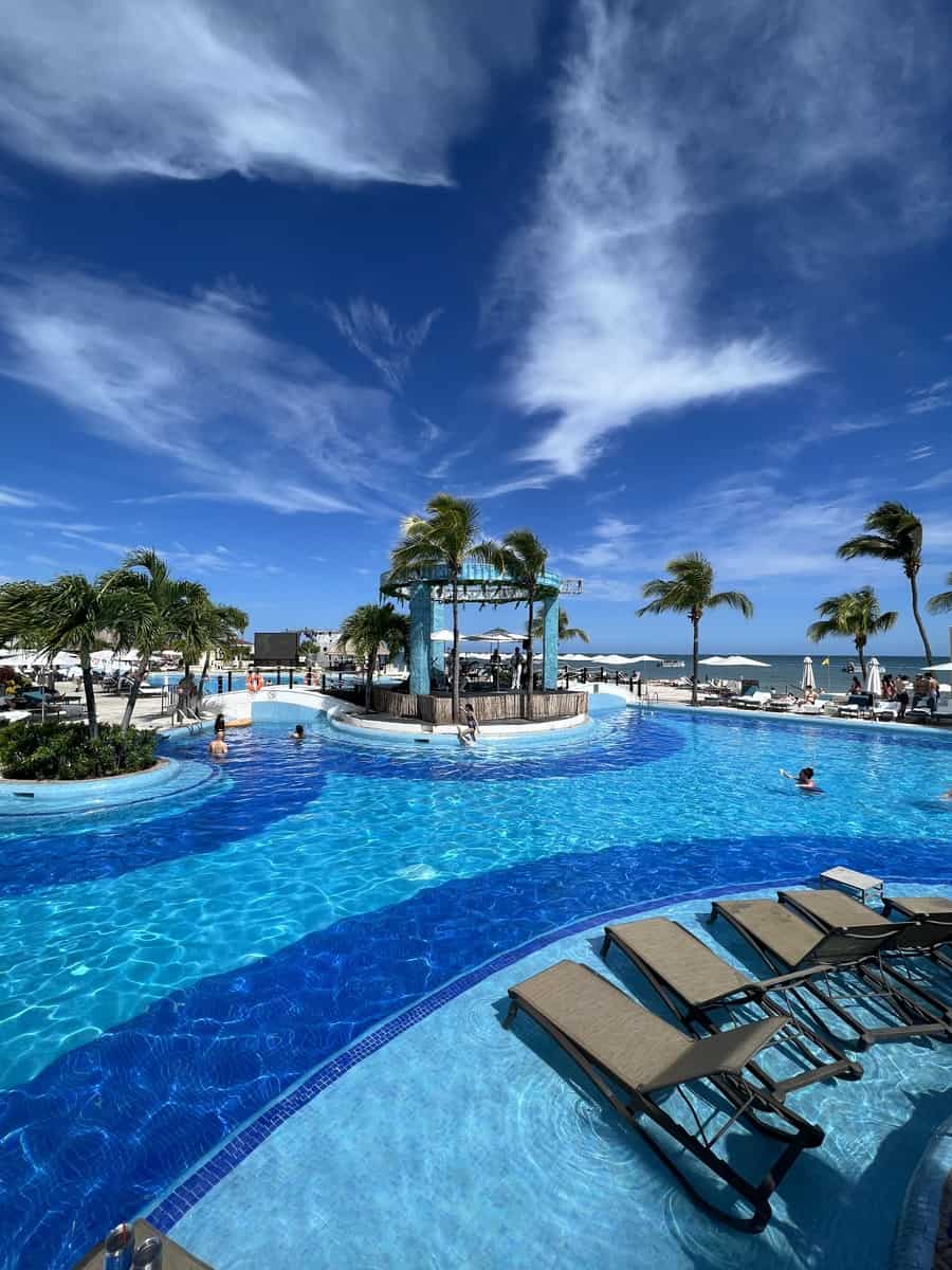 Large pool with chairs, palm trees, and the Caribbean sea in the background on a sunny day.