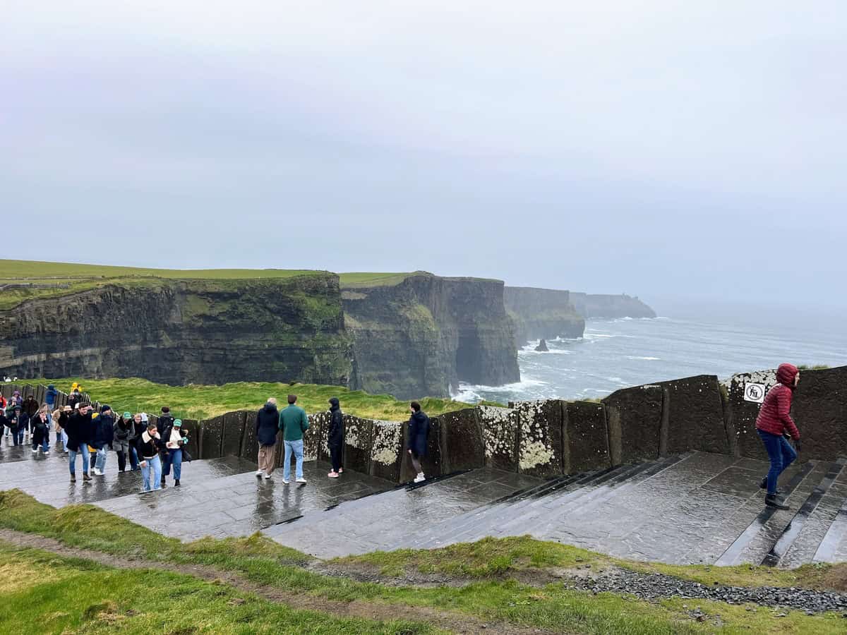 The image shows a group of tourists visiting the Cliffs of Moher in Ireland on a cloudy and misty day. The iconic cliffs, with their sheer rock faces and patches of green grass on top, stretch into the distance, meeting the turbulent sea below. The viewing area features wide stone steps and paths, where visitors, dressed in warm jackets and hats, are walking, taking photos, and enjoying the view.