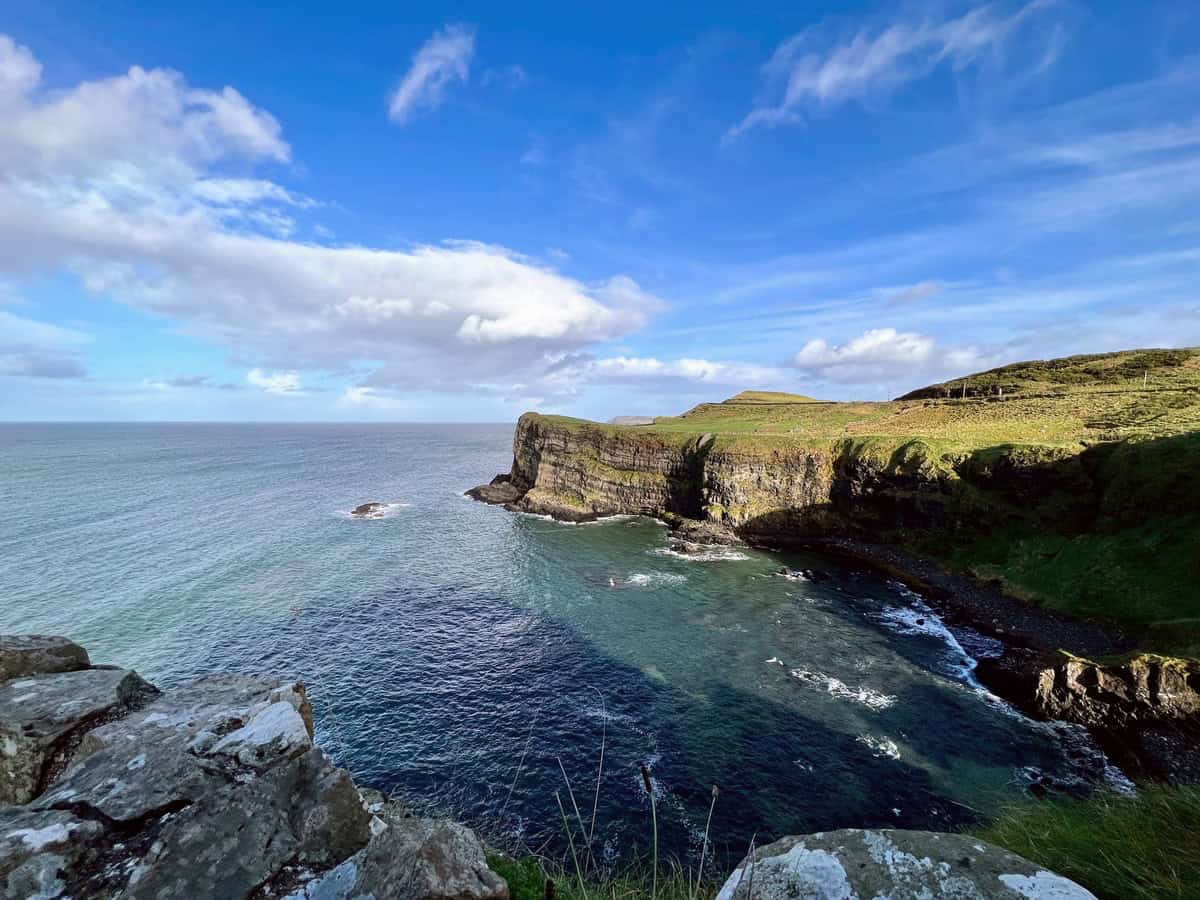 Northern Ireland scenic coastal landscape featuring a dramatic cliff jutting out into the ocean under a bright blue sky. The cliff, covered in green grass and rocky outcrops, descends steeply into dark, swirling waters below.
