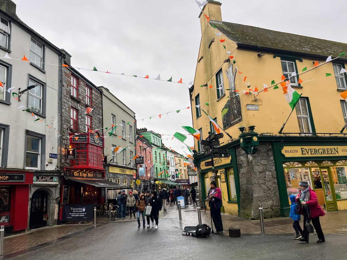 The image shows a lively street scene in Galway City, Ireland, adorned with colorful bunting in green, white, and orange, reminiscent of the Irish flag. The street is lined with various shops and pubs, each with vibrant facades. On the left, there is a pub called "The Kings Head," featuring a distinctive red exterior with a sign promoting Guinness. A street musician is performing near the corner.