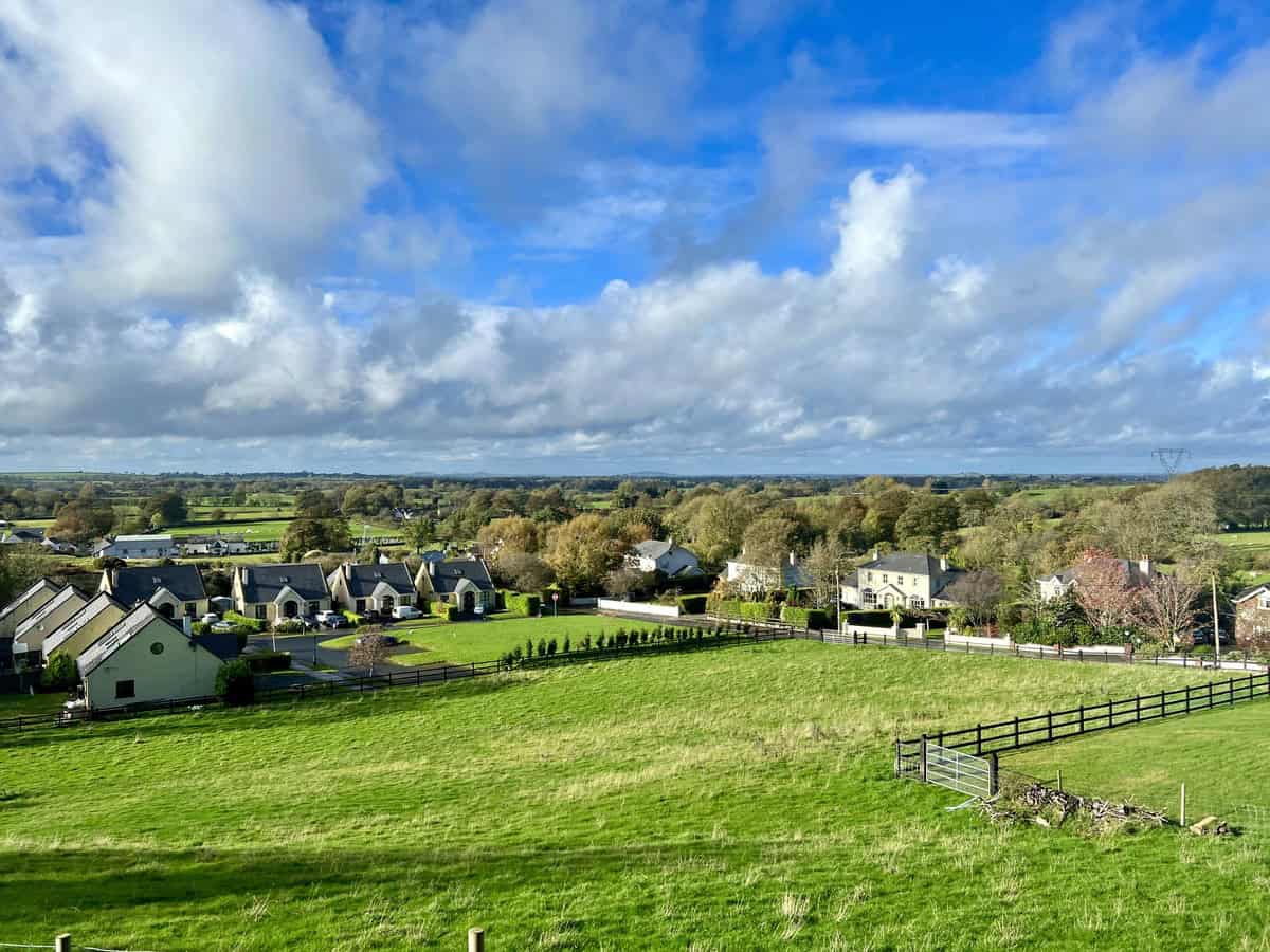 The image features the charming rural village, Hollywood, in County Wicklow, Ireland, set in a lush, green landscape. In the foreground, there is a large, open grassy field bordered by a wooden fence. The landscape extends into a patchwork of fields and woodlands under a vibrant blue sky dotted with fluffy white clouds.