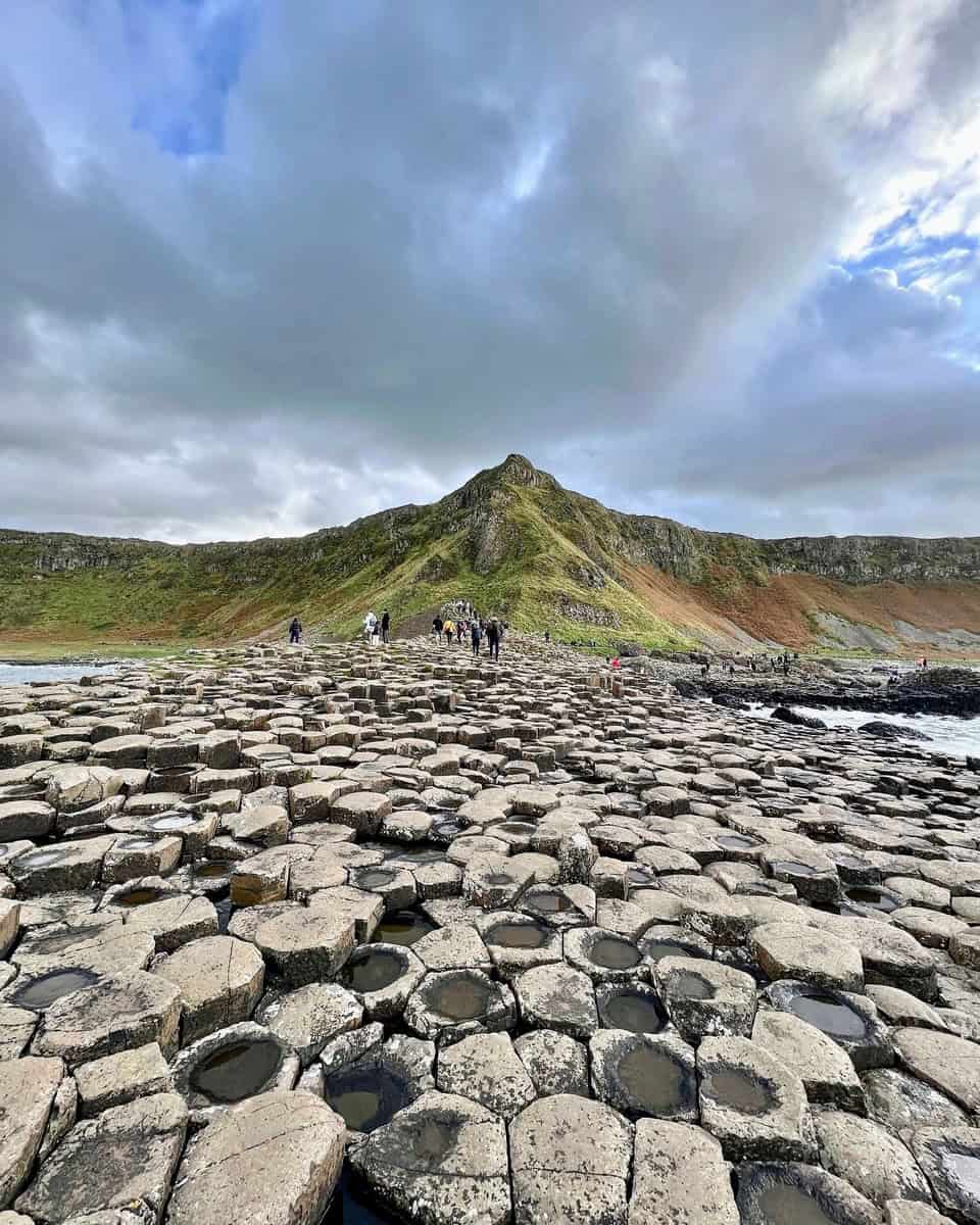 The image shows a group of people exploring the Giant's Causeway, a famous natural landmark on the coast of Northern Ireland. The terrain consists of numerous hexagonal basalt columns that create a unique, stepped formation, extending into the sea.