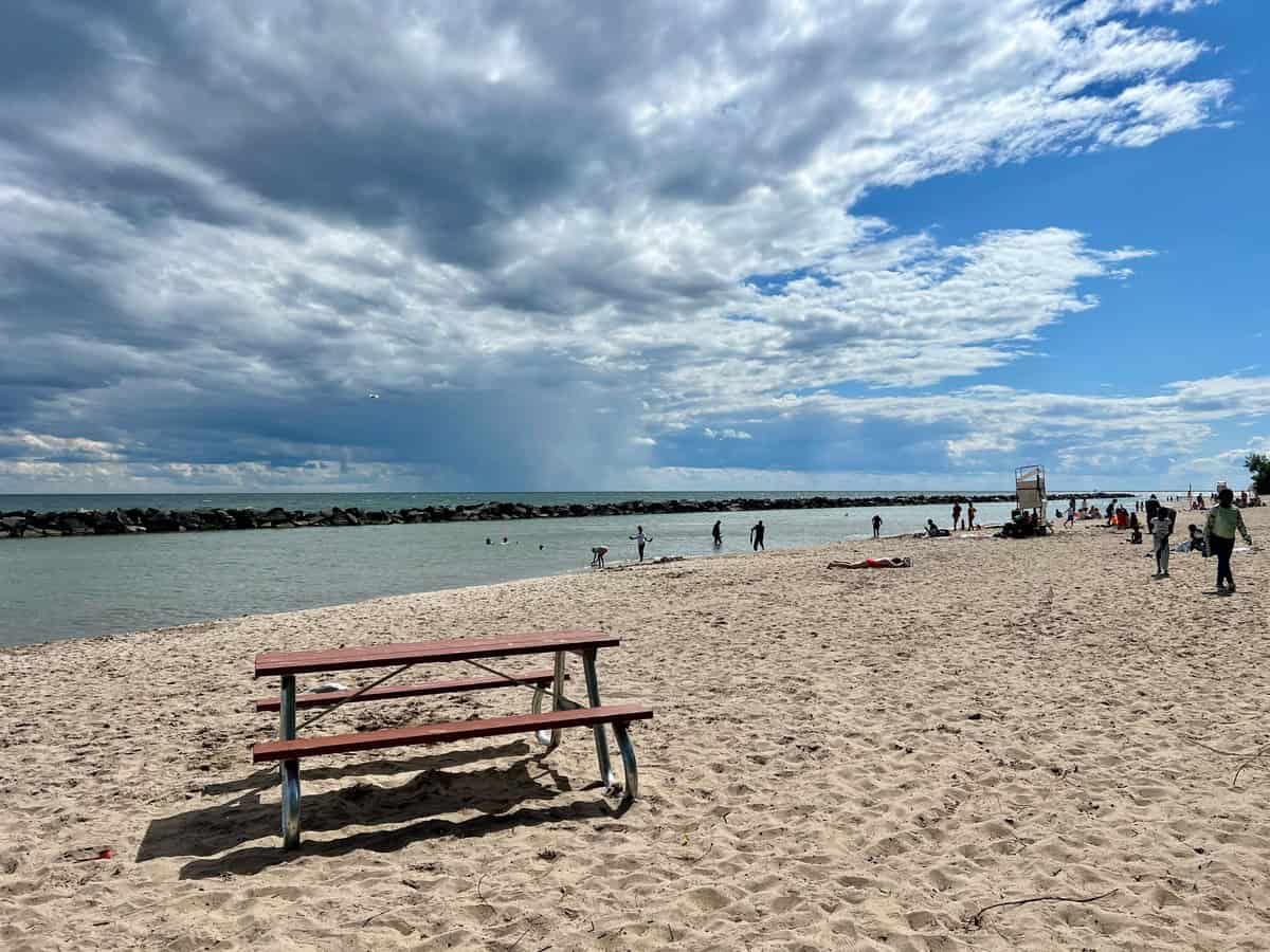 A calm beach scene with clear, shallow water gently rippling onto the sandy shore on the Centre Island Beach in Toronto. In the background, a rocky breakwater extends across the water under a sky filled with puffy clouds.