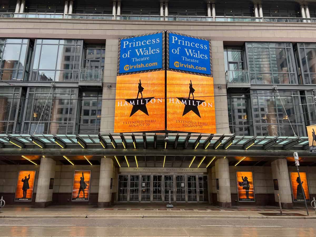 The exterior of the Princess of Wales Theatre in Toronto, prominently displaying advertisements for the musical Hamilton. The theater's modern glass facade and marquee lights are visible, with reflections on the wet sidewalk below.