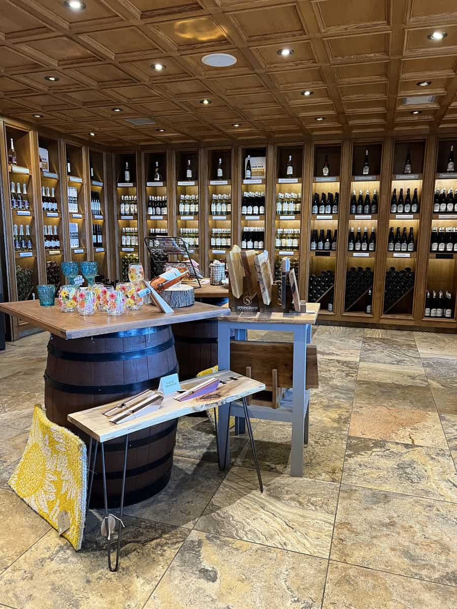 An elegantly designed wine store with wooden shelves filled with bottles of wine lining the walls. In the center, there are displays featuring wine accessories and decorative items placed on a barrel and a small table. The store has a warm and inviting ambiance with a tiled floor and a coffered ceiling.
