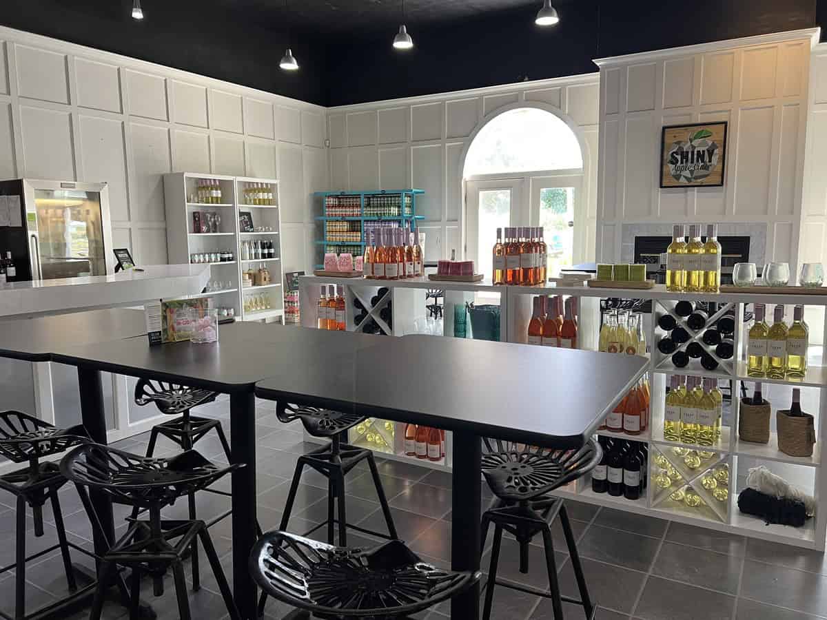 The interior of a bright and modern cider shop, featuring a clean, white aesthetic with bottles of cider and other products displayed on shelves. There are tall tables with bar stools in the center, and the space has a minimalist, stylish design.