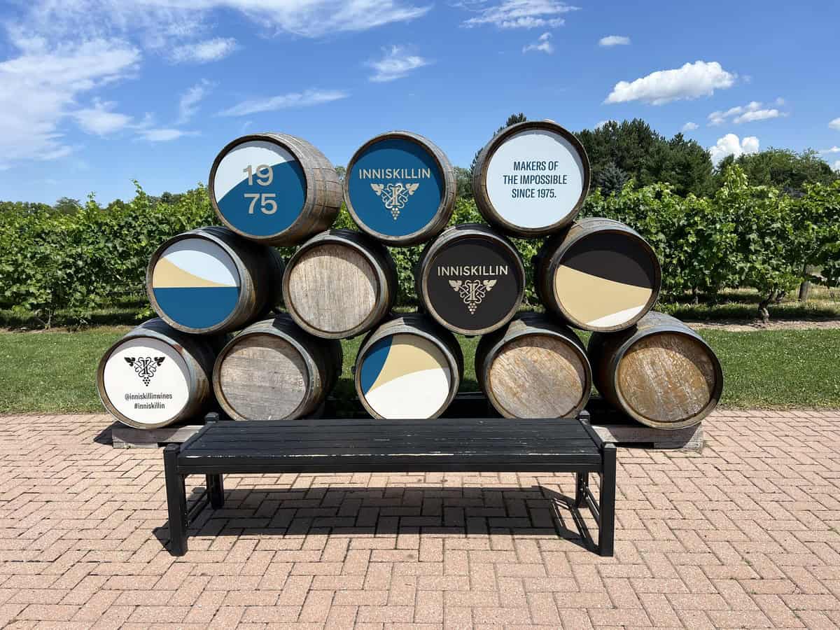 A display of nine wooden barrels stacked in a pyramid shape at one of the best Niagara on the Lake wineries, Inniskillin. The barrels feature logos and text including "1975" and "Makers of the Impossible since 1975." A black bench is in front, with a background of lush Niagara on the Lake vineyards under a blue sky, showcasing a top-rated winery in NOTL.
