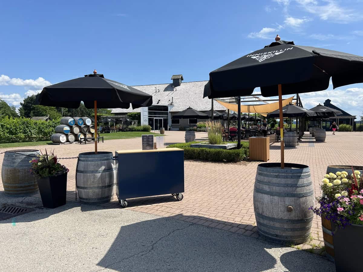 The outdoor seating area of Inniskillin Winery, featuring black umbrellas, wooden barrels, and a backdrop of vineyard rows under a bright blue sky, exemplifying one of the best Niagara wineries.