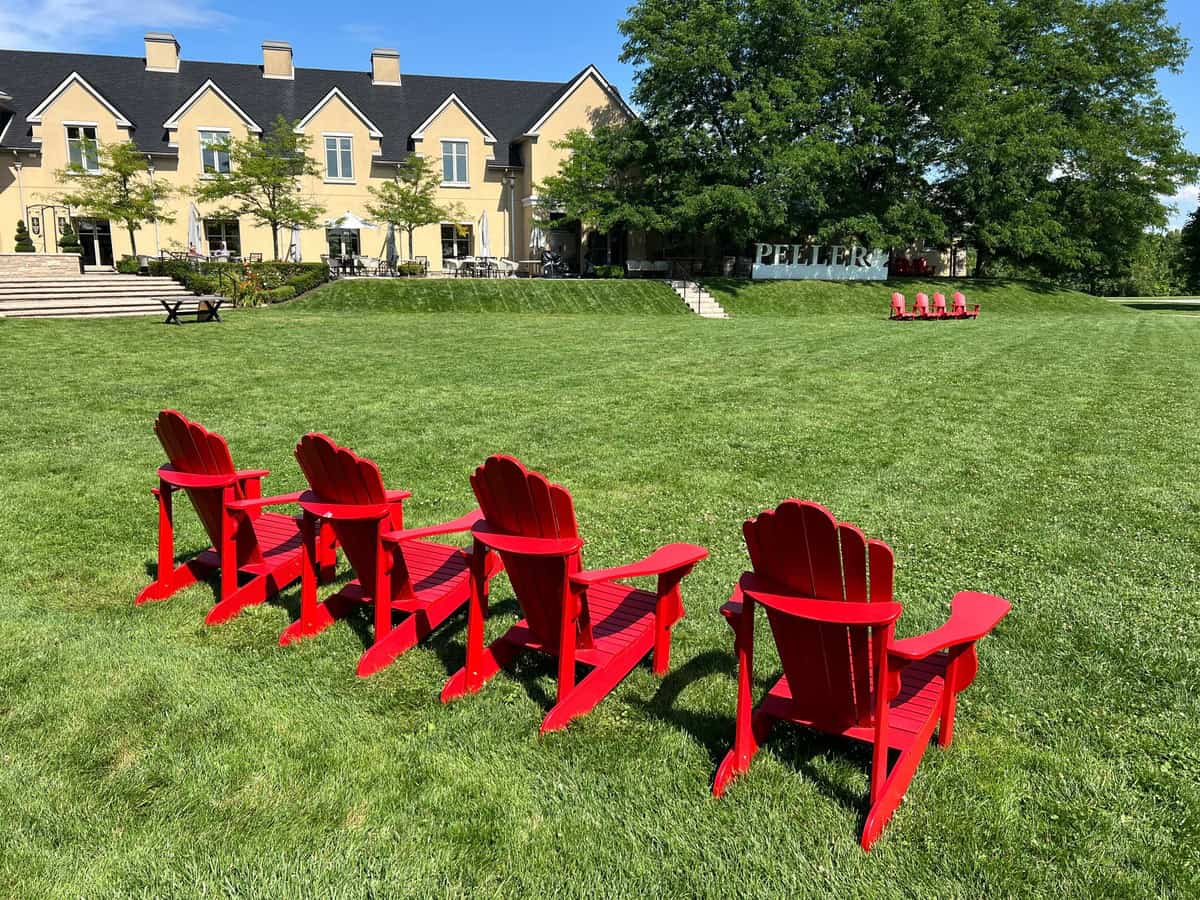 A row of red Adirondack chairs on a spacious lawn facing the elegant Peller Estates building, offering a serene view perfect for relaxing at one of the best Niagara on the Lake wineries.