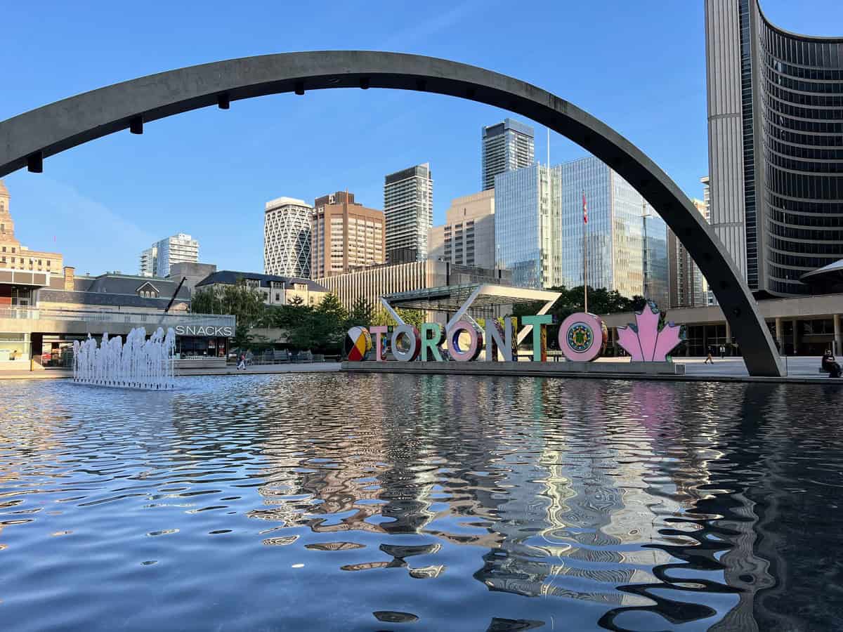 The iconic "Toronto" sign at Nathan Phillips Square is reflected in the calm waters of the square's fountain pool. The sign is framed by the large, curved concrete arch that spans the square, with the city’s modern skyscrapers and historic buildings in the background. Toronto's City Hall is behind the sign.