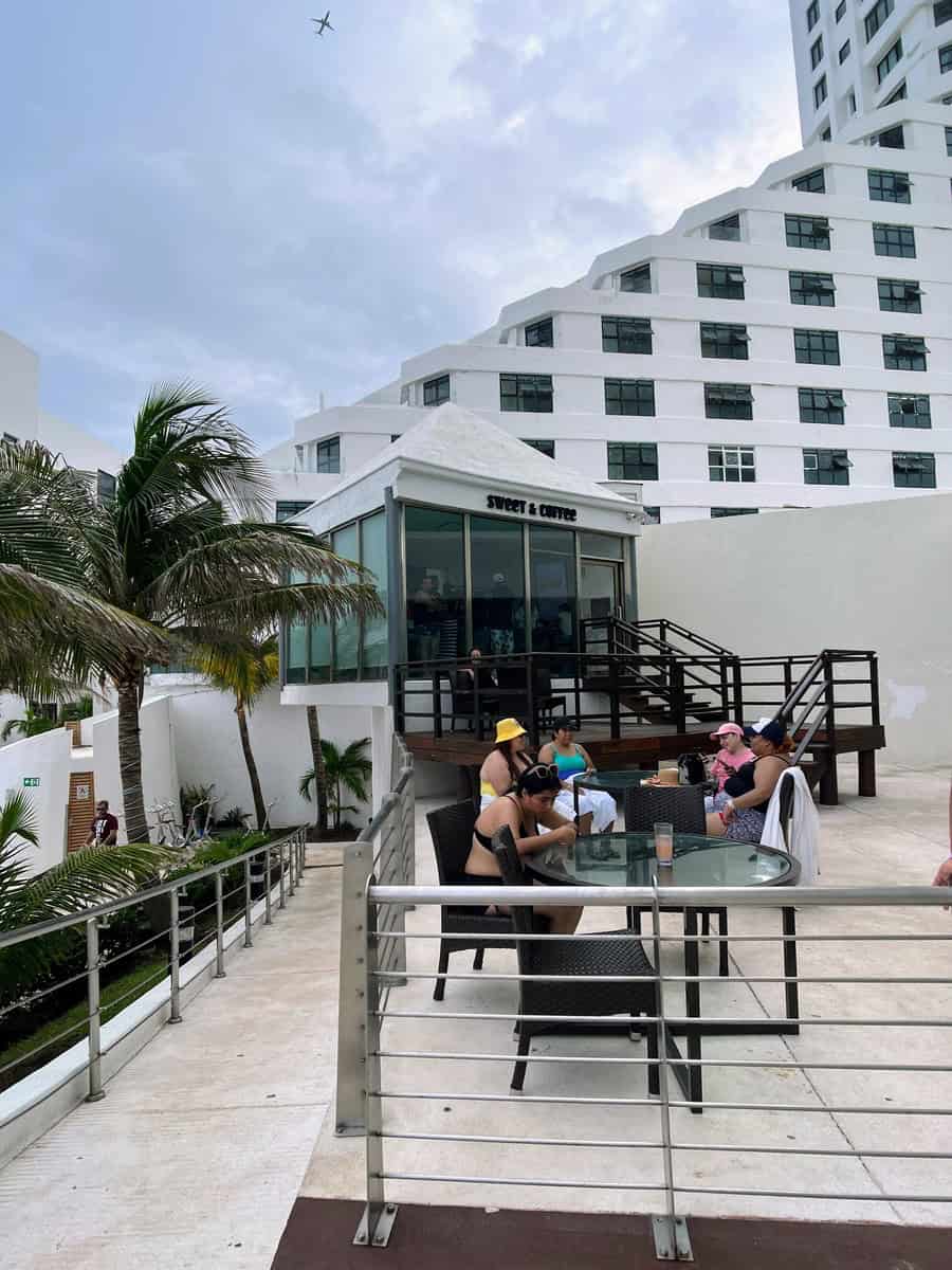 Outdoor seating area at the Sweet & Coffee shop in Beach Palace Resort, with guests relaxing at round glass tables. The modern, glass-walled café sits on an elevated platform, offering views of the surrounding area and a casual spot to enjoy refreshments.
