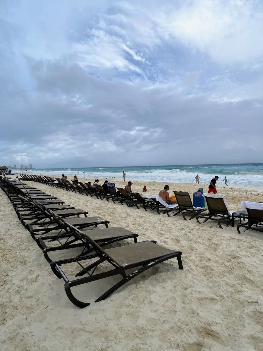 Rows of lounge chairs lined up on the white sandy beach at Beach Palace Resort, overlooking the turquoise waters of the Caribbean Sea. Guests relax by the shore, enjoying the tropical beach setting under partly cloudy skies.