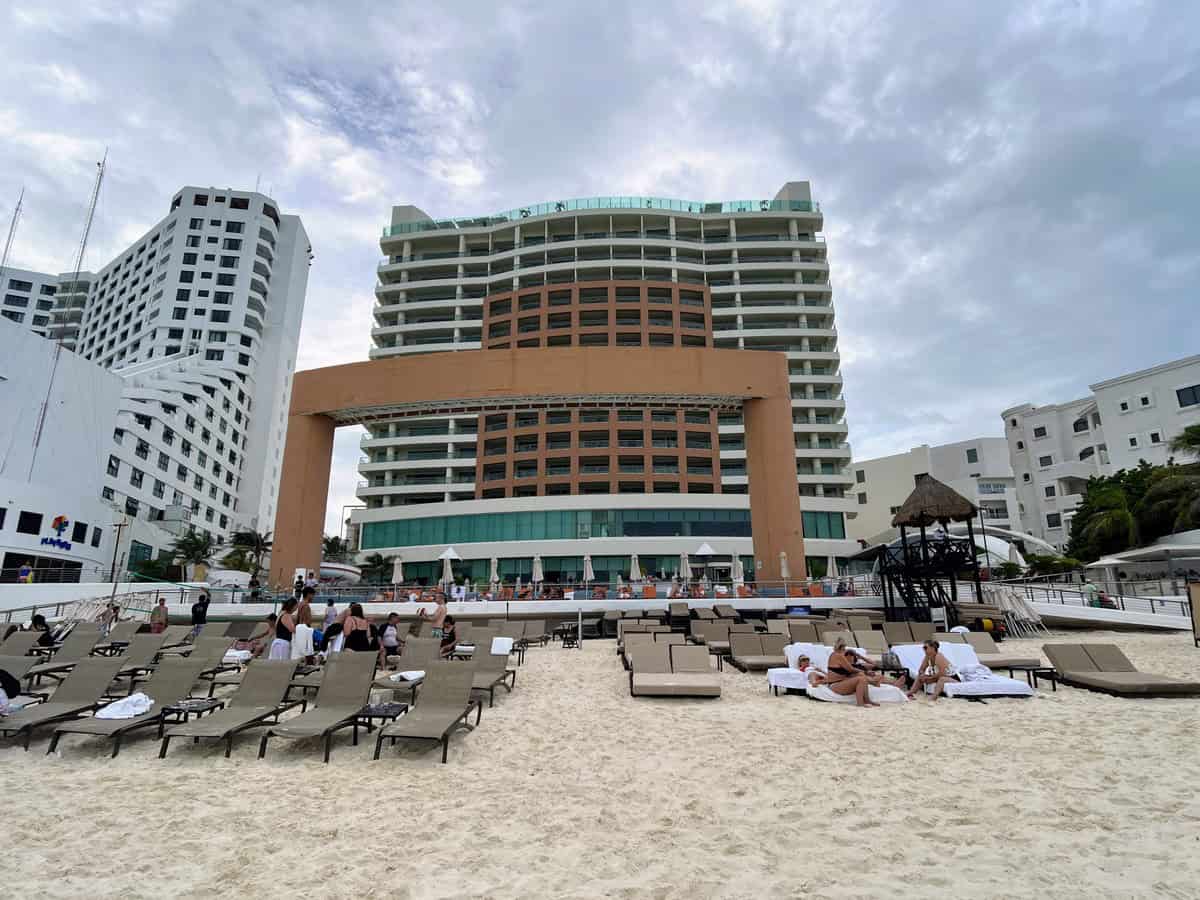 Beachfront view of the Beach Palace Resort in Cancun, featuring a modern high-rise building with sun loungers arranged on the sandy beach. Guests relax on the beach, enjoying direct access to the ocean and resort amenities.