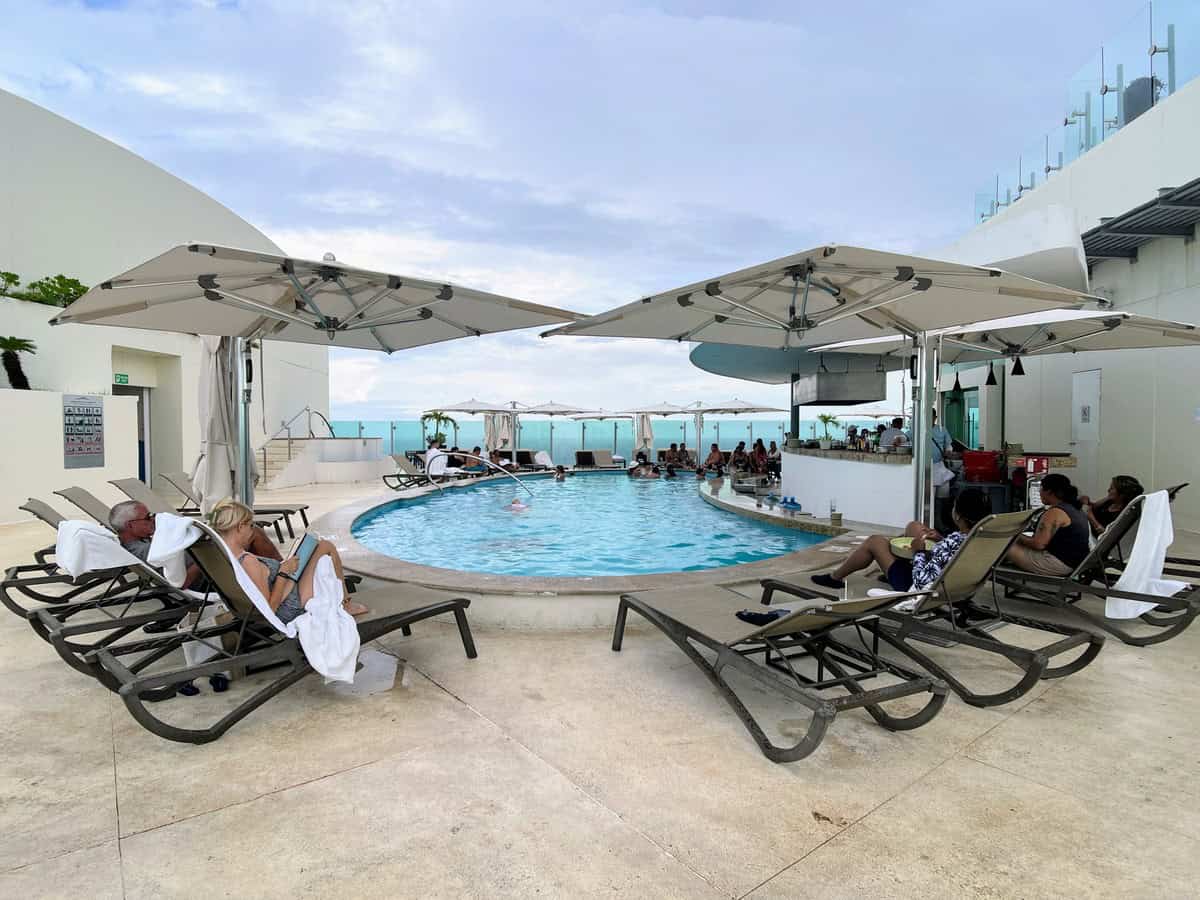 Rooftop pool at Beach Palace Resort in Cancun, surrounded by umbrellas and sun loungers. Guests relax by the pool, enjoying the elevated views of the turquoise ocean under a clear sky.