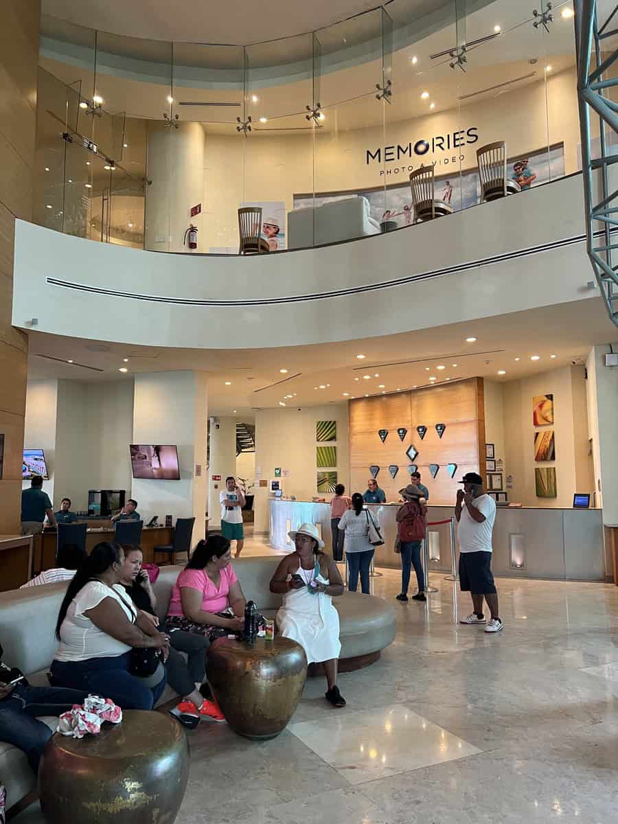 Spacious lobby at Beach Palace Resort featuring comfortable seating areas, modern decor, and guests relaxing and checking in. The open, two-story design includes a Memories photo studio on the mezzanine level.