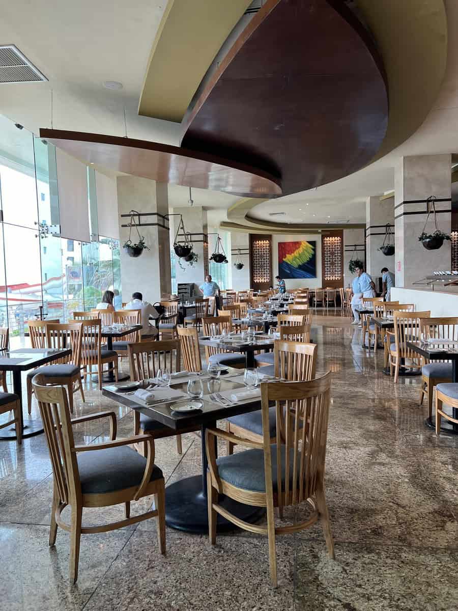 Modern dining area inside a restaurant at Beach Palace Resort, featuring sleek wooden chairs, glass-topped tables, and large windows with plenty of natural light. The decor includes hanging planters and a unique, curved ceiling element that adds to the stylish ambiance.