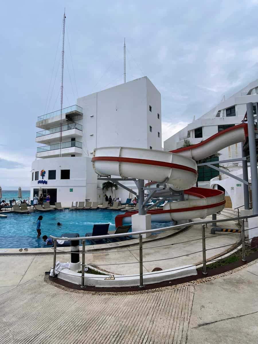 Pool area at Beach Palace Resort featuring a fun water slide that loops into the pool. Families and children enjoy swimming and playing around the slide, while lounge chairs are arranged poolside for relaxation.