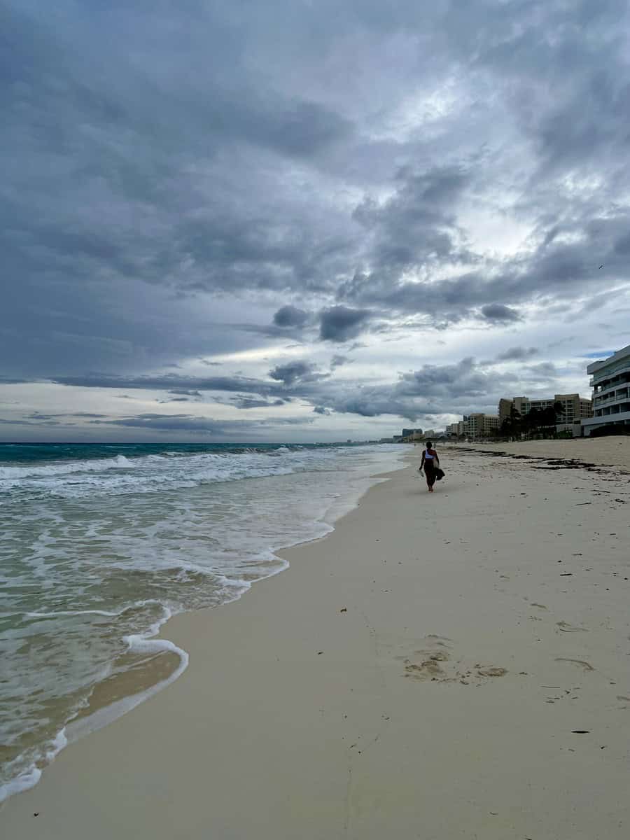 Expansive view of the beach at Moon Palace Cancun, with gentle waves rolling onto the soft, sandy shore. A lone person walks along the coastline, while overcast skies create a serene, tranquil atmosphere.