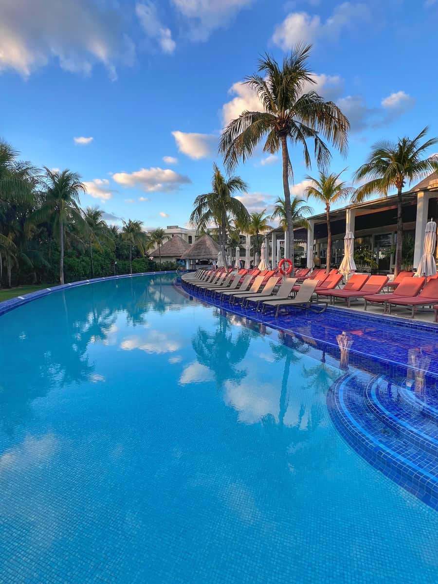 Crystal-clear, blue-tiled pool at Moon Palace Cancun The Grand, surrounded by rows of red lounge chairs and tall palm trees. The serene pool reflects the vibrant sky and palm trees, creating a tropical oasis perfect for relaxation.