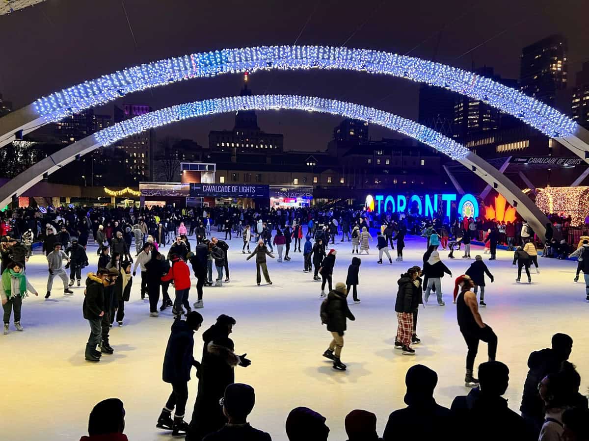 Nathan Phillips Square during the Cavalcade of Lights Christmas activity, featuring a large crowd ice skating under brightly lit arches with the Toronto sign illuminated in the background and city lights glowing above.