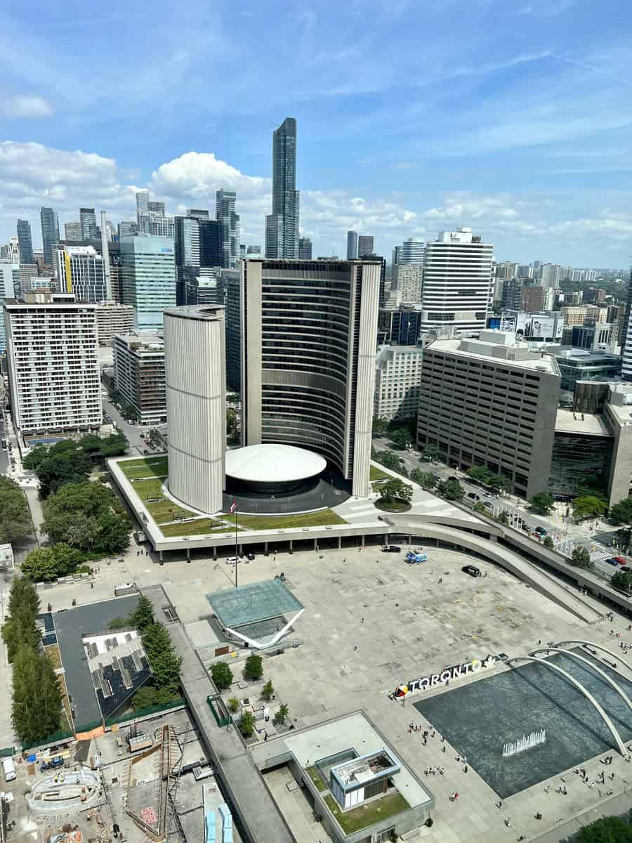 Aerial view of Nathan Phillips Square during the day, showcasing the Toronto sign downtown, the modern City Hall, surrounding skyscrapers, and a few people walking through the square.