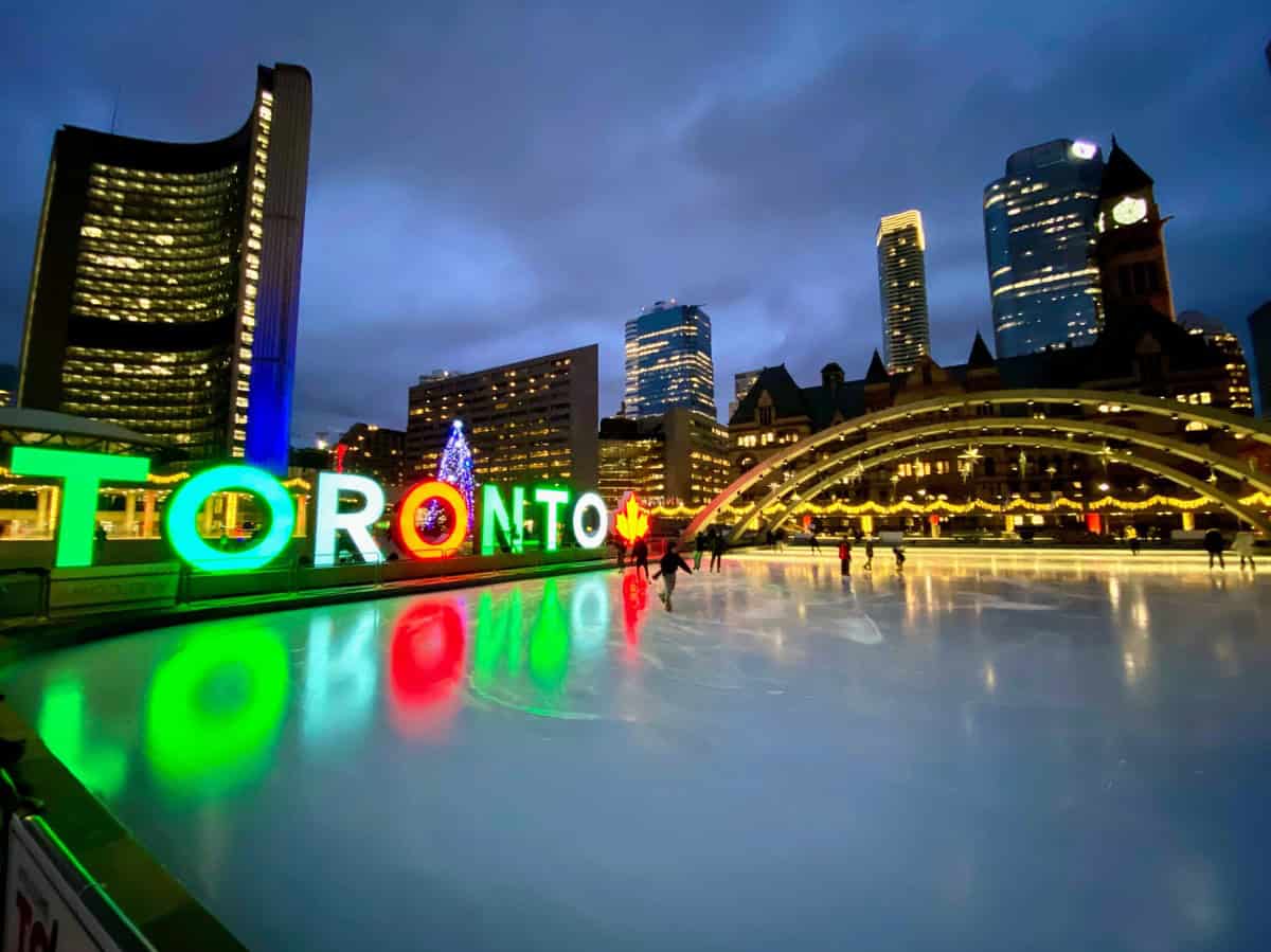 Nathan Phillips Square at night during winter, featuring the brightly lit Toronto sign in green, red, and white colors reflecting on the ice skating rink, with a lit Christmas tree and illuminated buildings in the background.