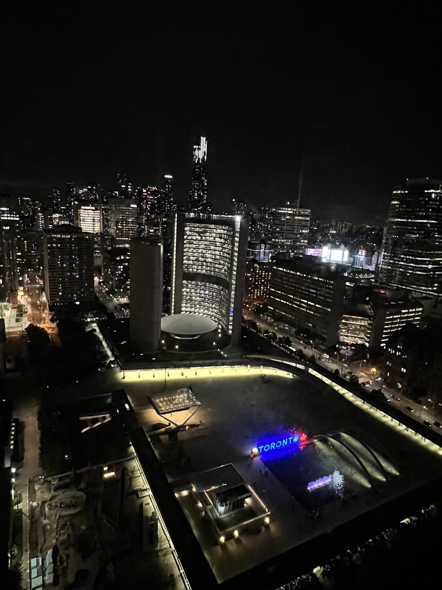 A nighttime aerial view of Nathan Phillips Square, with the brightly lit Toronto sign visible amidst the surrounding skyscrapers and Toronto city hall.