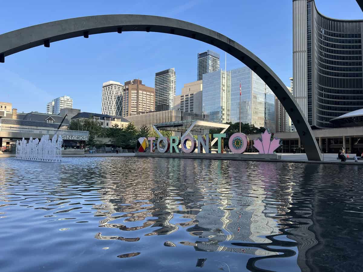 The Toronto sign downtown at Nathan Phillips Square in summer, reflected in the water of the fountain, surrounded by Toronto skyscrapers and bathed in warm sunlight.