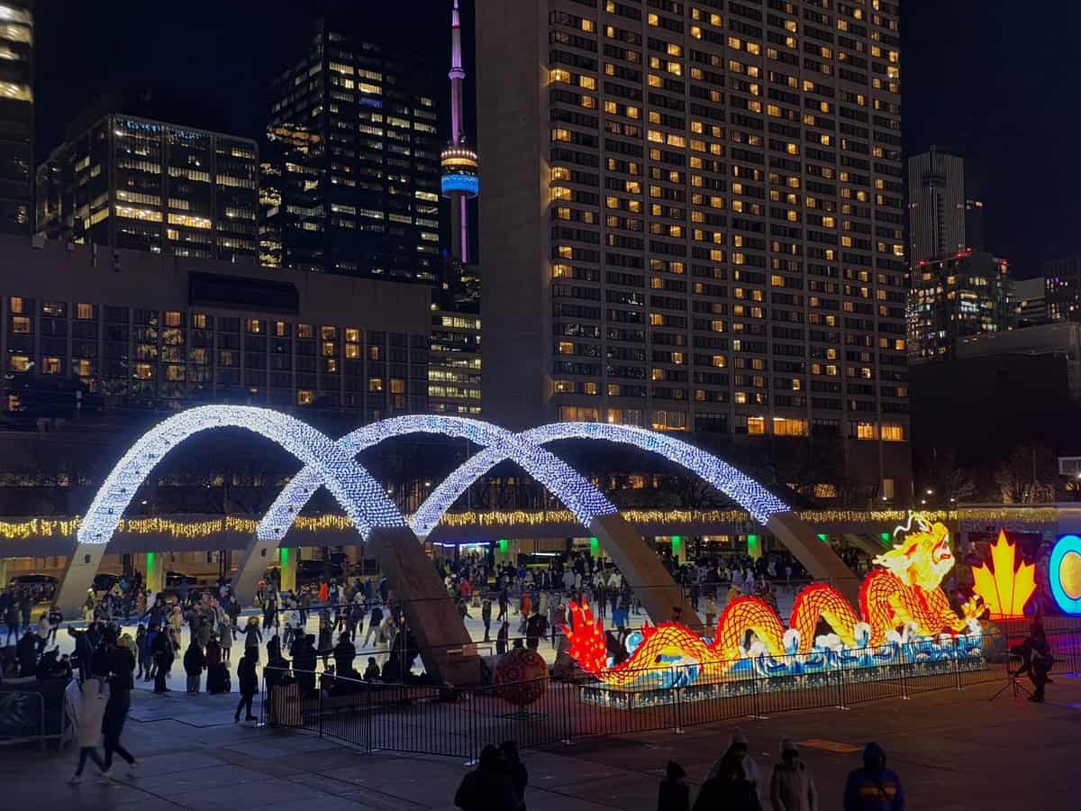 A vibrant nighttime scene at Nathan Phillips Square in Toronto featuring glowing white arches, a bright red and yellow illuminated dragon decoration, and the CN Tower lit up in purple and orange in the background. Ice skaters glide across the rink surrounded by illuminated buildings during the holidays.