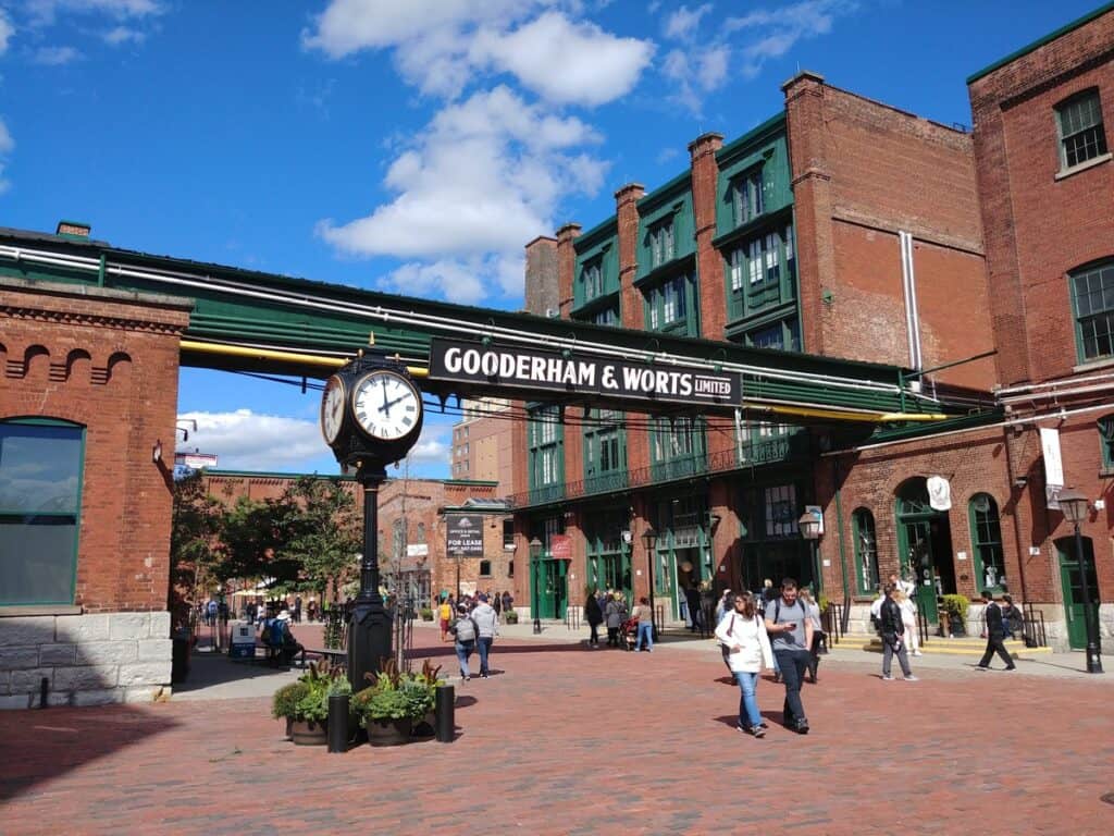 The historic Toronto Distillery District on a sunny fall day, featuring the Gooderham & Worts sign, a classic black clock, and red brick buildings with green accents. Visitors stroll along the cobblestone streets, enjoying the vibrant atmosphere.