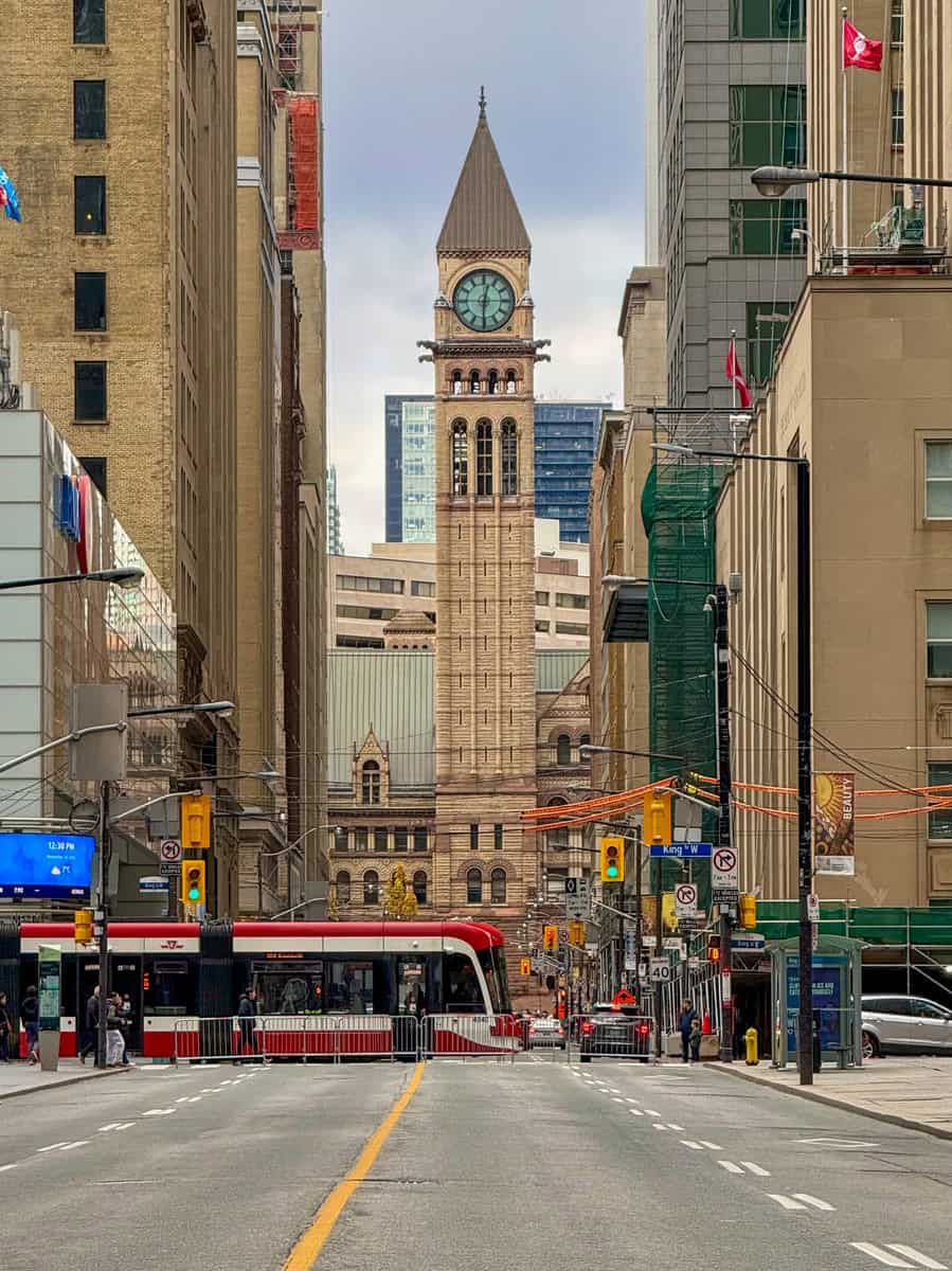 A downtown Toronto street scene with the historic Old City Hall clock tower standing tall in the background. A modern red and white TTC streetcar is stopped at an intersection, with pedestrians crossing and traffic signals glowing. The surrounding buildings display a mix of old and new architecture, with construction scaffolding and Canadian flags adding to the urban atmosphere.