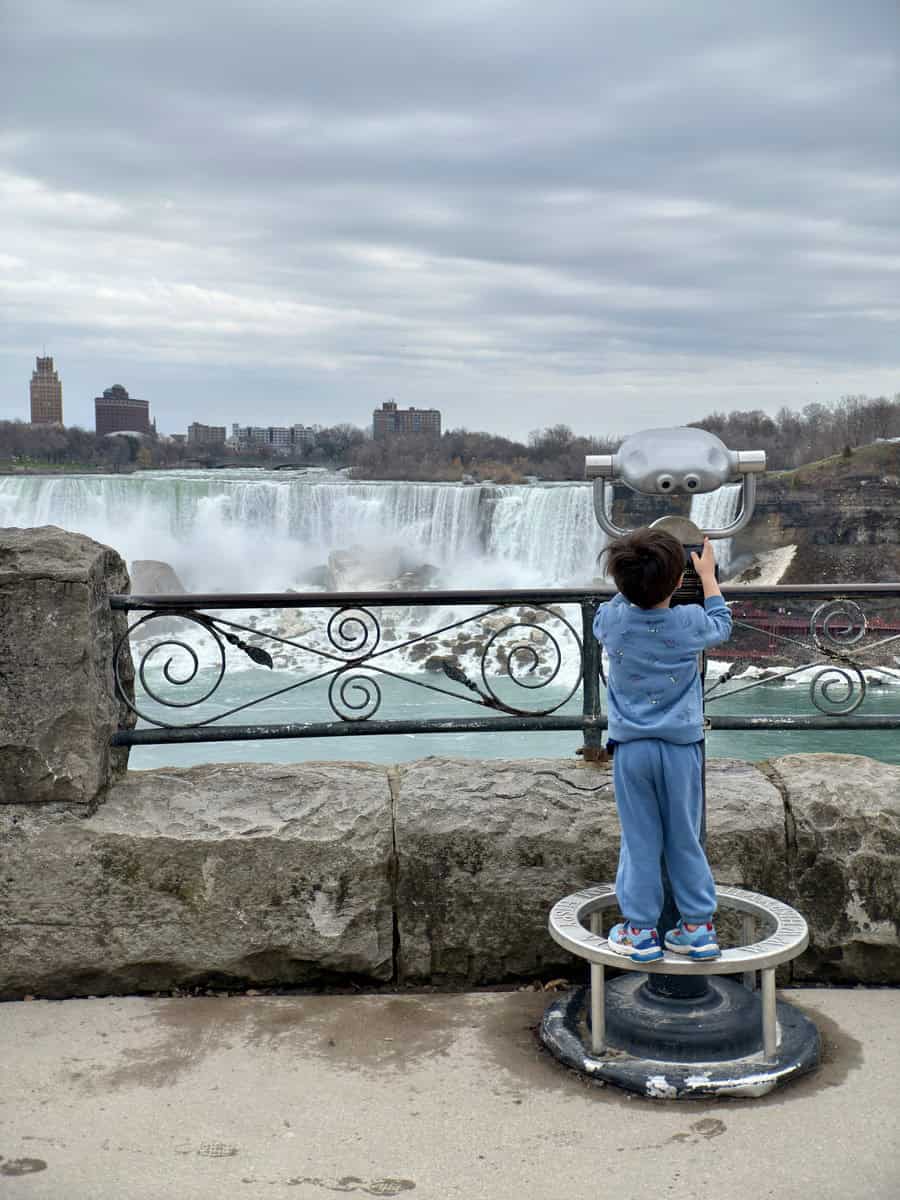 A young child in blue clothing stands on a metal viewing stand, peering through binoculars at the misty American Falls with city buildings in the distance. A sweet moment from a family-friendly Niagara Falls itinerary.