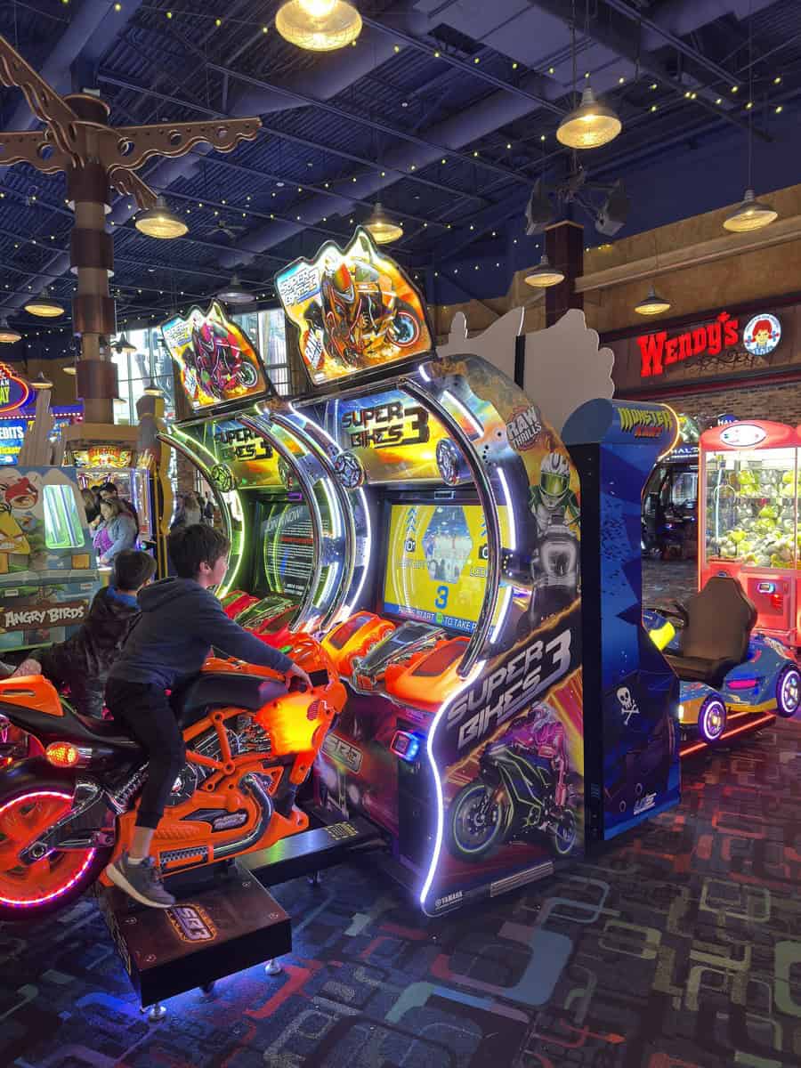 A child plays on a glowing orange motorcycle arcade game inside a lively gaming center with neon lights and a Wendy’s sign in the background. A fun indoor activity spot highlighting things to do in Niagara Falls with kids.
