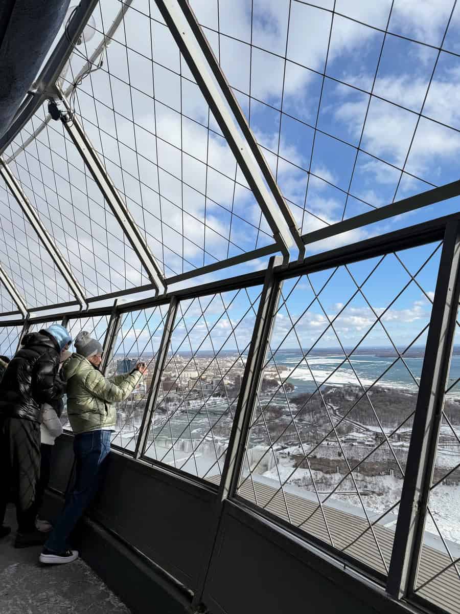 Visitors bundled in winter jackets lean against the fenced windows of the Skylon Tower outdoor observation deck, capturing snowy views of Niagara Falls and the icy river below.