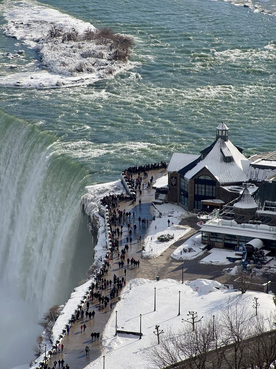 Aerial view from the Skylon Tower showing crowds gathered at Table Rock near the edge of Horseshoe Falls, with snow-covered walkways and a frozen river creating a stunning winter scene, perfect for a Skylon Tower review.