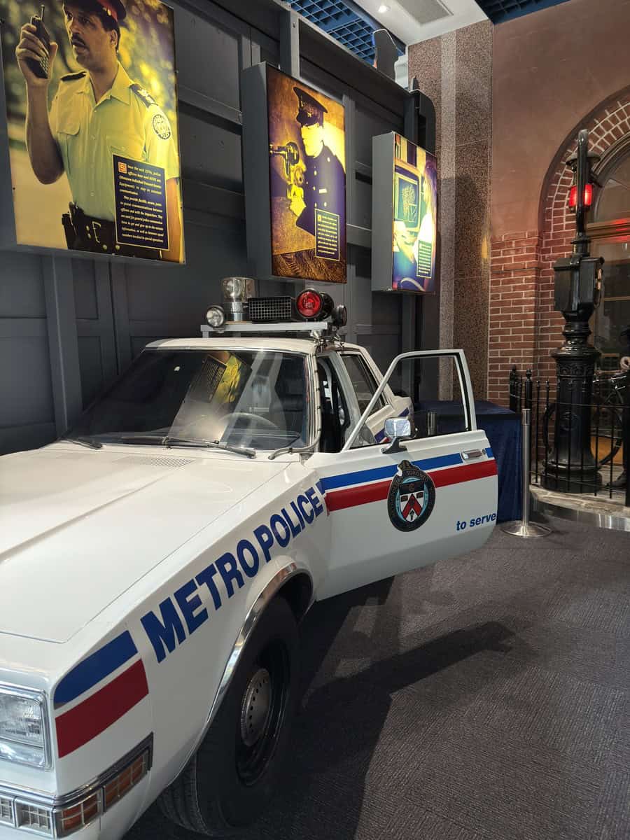 A white retro Toronto Metro Police car with red and blue striping is displayed indoors with the driver’s door open, showcasing the Toronto Police Service badge on the side. Behind it, illuminated panels feature historic images of police officers using communication technology, adding context to the exhibit.