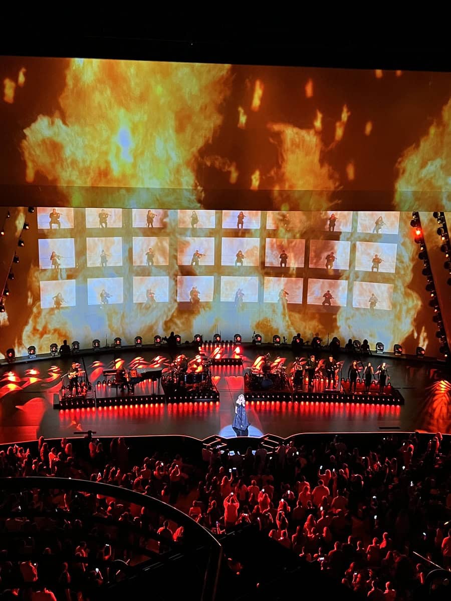 Adele stands center stage in Las Vegas, surrounded by red and orange lighting and a backdrop filled with flames and silhouetted performers in video panels. The dramatic fire-themed visuals and moody lighting create an intense, theatrical moment.