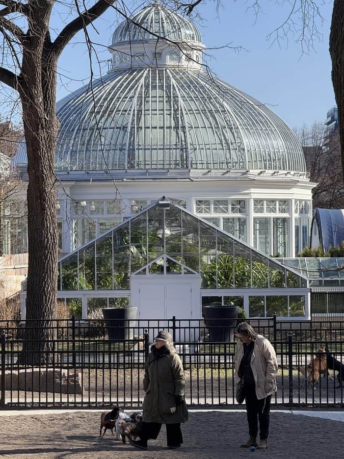 Two people walk small dogs near a fenced dog area in front of the grand glass-domed conservatory at Allan Gardens in Toronto on a crisp, sunny day.