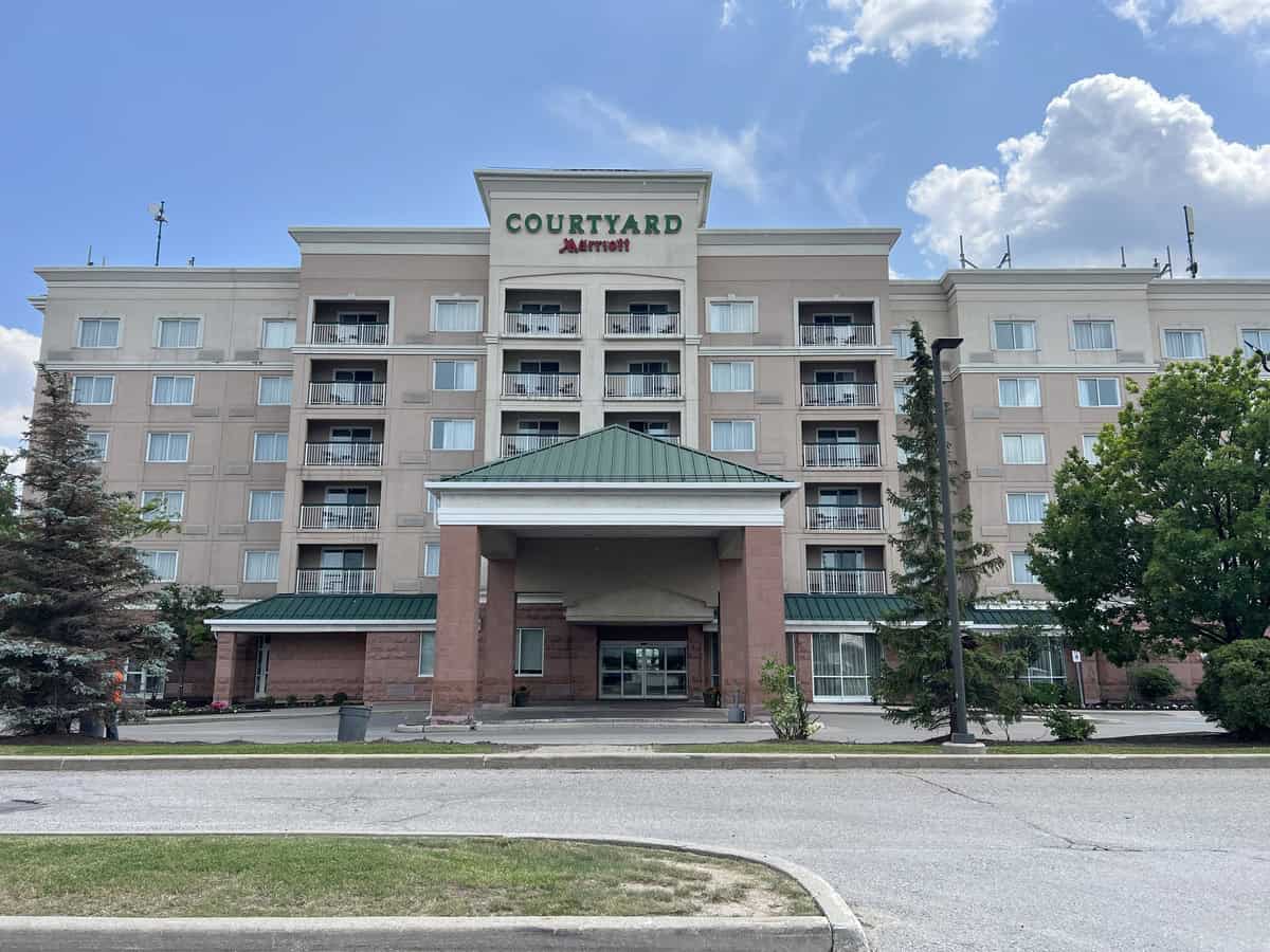 Front view of the Courtyard by Marriott hotel in Vaughan, Ontario, showing its multi-storey beige and pink exterior with balconies, a green metal roof, and a covered entrance under a partly cloudy sky.