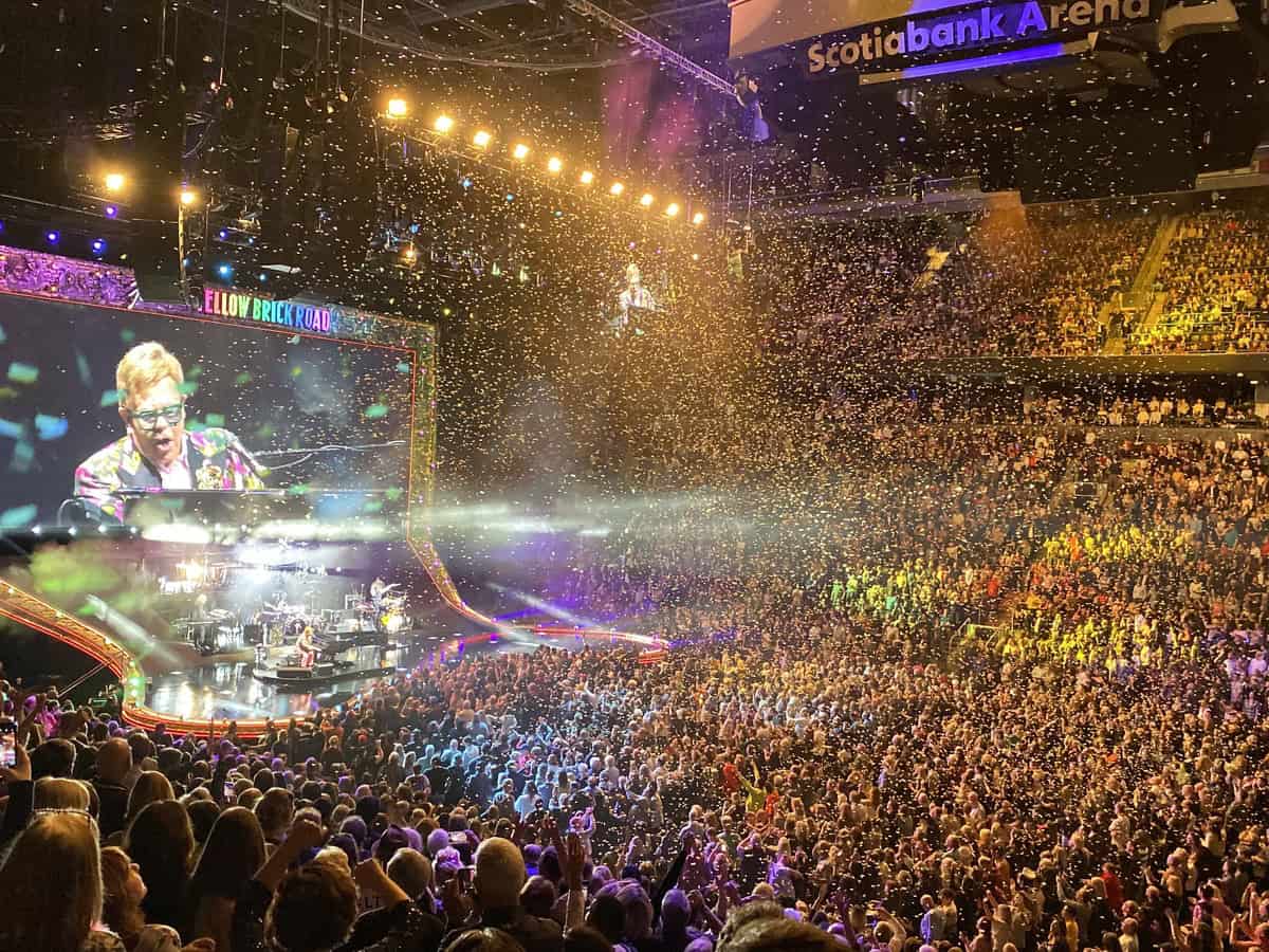 Elton John performs at the Scotiabank Arena in Toronto during his Farewell Yellow Brick Road tour, wearing a colorful jacket at his piano under golden confetti. A massive screen behind him shows his image as rainbow-colored stage lights and a cheering crowd fill the arena.